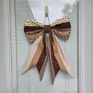 May include: A decorative fabric bow featuring a patchwork design with various brown, tan, and cream patterns including floral, striped, and geometric prints. The bow has two large top loops and two long, pointed tails hanging down from a central knot.