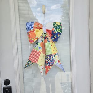 May include: A large decorative bow made from a patchwork of colorful cotton fabrics featuring patterns of oranges, strawberries, lemons, and gingham checks in shades of yellow, orange, red, green, and blue, suspended by a white cord loop.