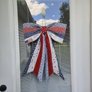 May include: A decorative fabric bow featuring a patriotic theme with red, white, and blue colors. The bow displays various patterns including stars, stripes, small bow ties, and a small American flag graphic on a white background with text.