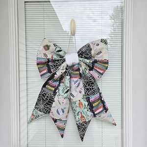 May include: A large fabric bow featuring a pattern of potion bottles, stacked books, and spider webs in black, white, green, and orange. The bow has a solid white center knot and two long, pointed tails hanging down, suspended by a small beige hook.