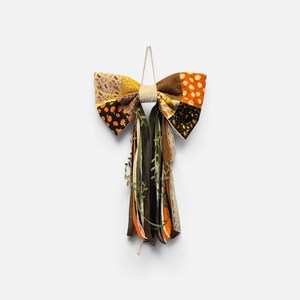 May include: A decorative bow made from patchwork fabric featuring autumn-themed patterns of pumpkins and leaves in orange, brown, black, and cream. The bow includes long, hanging fabric ribbons in solid colors, lace, and faux greenery vines.