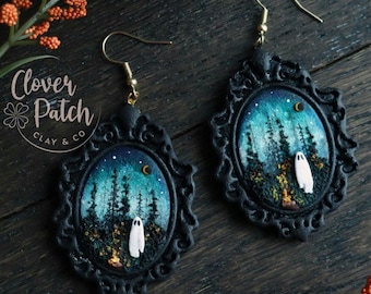 May include: A pair of black framed earrings with a spooky forest scene. Each earring features a night sky with a crescent moon, trees, and a white ghost figure. The earrings have gold-colored hooks and the text "Clover Patch Clay & Co".