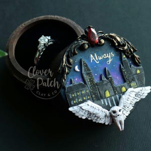 Peut inclure: Une boîte à bague en bois foncé avec un couvercle en céramique noir et blanc représentant un hibou des neiges aux ailes déployées, une lune croissante et une ville avec le texte "Always".