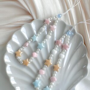 May include: Two beaded phone charms with star, heart, and seashell charms in pastel colors of pink, blue, and yellow. The charms are strung on a white cord with pearl beads. The charms are displayed on a white seashell-shaped dish.