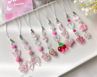 May include: Eight pink and white phone charms with different designs, including hearts, butterflies, flowers, strawberries, and cherries. The charms are strung on white cords and have silver metal clasps. The charms are displayed on a white surface with a pink and white card that says "Tinna Studio" in the background.