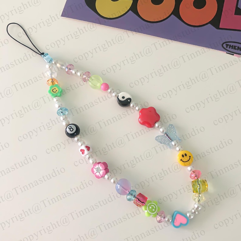 Pode incluir: Um amuleto de telefone de contas brancas com pingentes coloridos, incluindo um rosto sorridente, uma borboleta, um s&iacute;mbolo yin yang, uma flor vermelha, uma flor verde, um cora&ccedil;&atilde;o e uma bola de bilhar 8.
