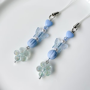 May include: Two blue and white butterfly phone charms with clear flower accents. Each charm features a blue heart, a blue bead, a white pearl, and a clear butterfly with blue wings.