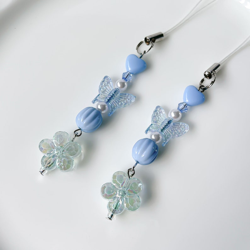 May include: Two blue and white butterfly phone charms with clear flower accents. Each charm features a blue heart, a blue bead, a white pearl, and a clear butterfly with blue wings.