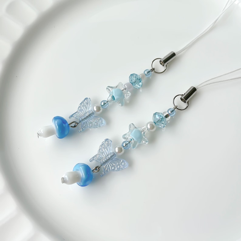 May include: Two blue and white phone charms with a mushroom, butterfly, and star design. The charms are made with beads and have a clear string.