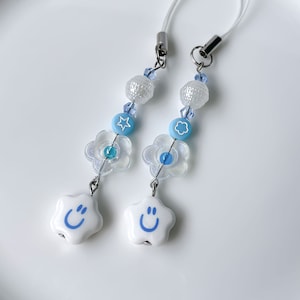 May include: Two white star-shaped charms with blue smiley faces, each attached to a blue and white beaded phone charm with a flower charm and a white bead.