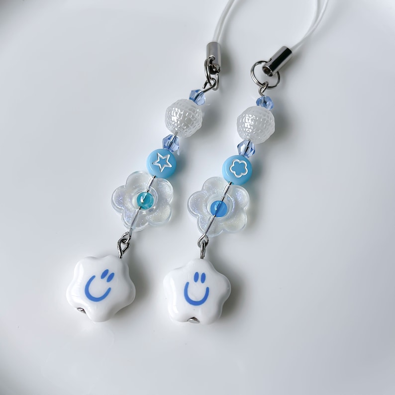 May include: Two white star-shaped charms with blue smiley faces, each attached to a blue and white beaded phone charm with a flower charm and a white bead.