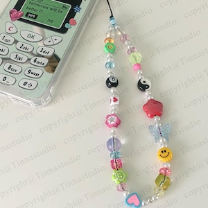 Pode incluir: Um amuleto de telefone de contas coloridas com uma variedade de pingentes, incluindo um rosto sorridente, uma borboleta, um s&iacute;mbolo yin yang, uma estrela vermelha, uma flor, um c&iacute;rculo verde e uma bola de bilhar preta e branca. O amuleto est&aacute; preso a um cord&atilde;o branco.