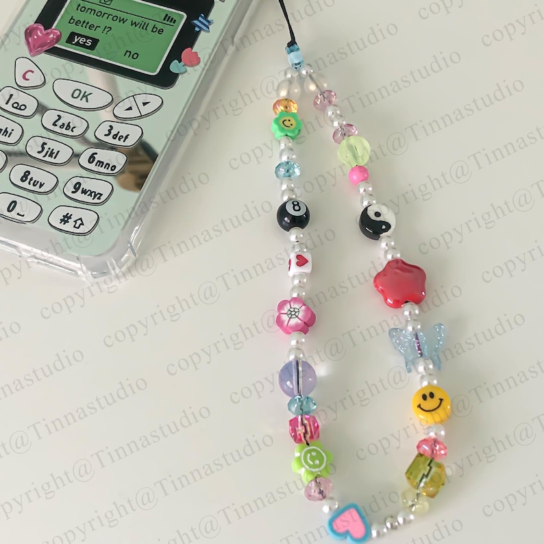 Pode incluir: Um amuleto de telefone de contas coloridas com uma variedade de pingentes, incluindo um rosto sorridente, uma borboleta, um s&iacute;mbolo yin yang, uma estrela vermelha, uma flor, um c&iacute;rculo verde e uma bola de bilhar preta e branca. O amuleto est&aacute; preso a um cord&atilde;o branco.