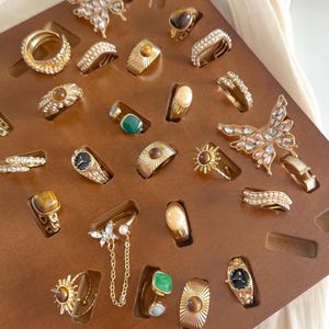 Könnte beinhalten: Eine Sammlung goldfarbener Ringe, präsentiert auf einem braunen Holzorganizer. Die Ringe weisen verschiedene Designs auf, darunter Edelsteine, Schmetterlingsakzente und aufwendige Details. Einige Ringe haben ein Zifferblatt-Design. Der Organizer steht vor einem weichen, cremefarbenen Hintergrund.