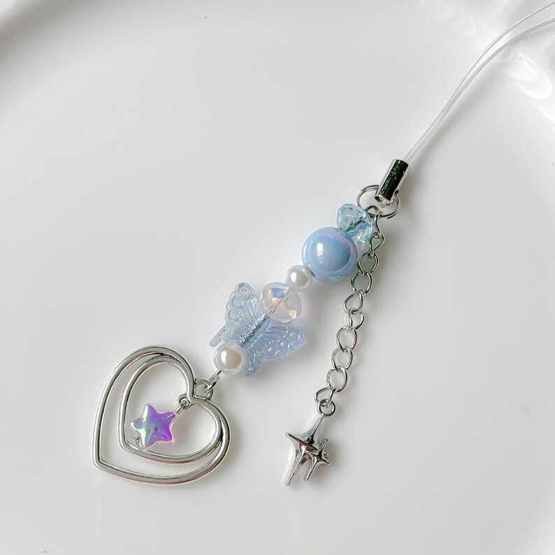 May include: A light blue and silver phone charm with a butterfly, heart, and star design. The charm is attached to a clear string.