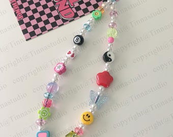 May include: A colorful phone charm with a variety of beads, including a smiley face, a butterfly, a star, a yin yang symbol, a flower, a heart, and a 8-ball. The charm is strung on a white pearl chain.