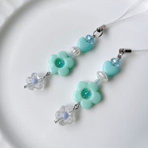 May include: Two turquoise blue phone charms with flower and heart beads. Each charm has a clear flower bead with a blue center and a turquoise blue heart bead with a clear rhinestone center.