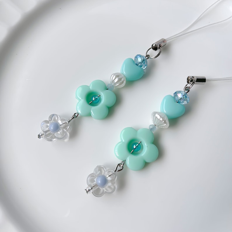May include: Two turquoise blue phone charms with flower and heart beads. Each charm has a clear flower bead with a blue center and a turquoise blue heart bead with a clear rhinestone center.