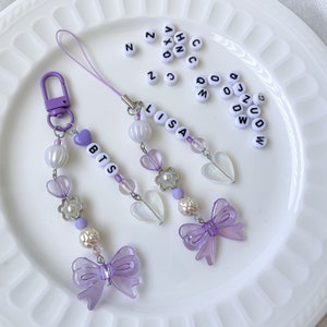Puede incluir: Tres abalorios para teléfono de color morado y blanco con los nombres "BTS", "LISA" y una sola letra "S". Cada abalorio tiene un lazo morado, cuentas blancas y una cuenta en forma de corazón.