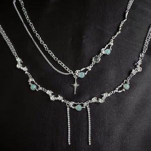 May include: A layered silver necklace set against a black background. The necklaces feature delicate chains, sparkling accents, and small, round, light blue beads. One necklace has a pendant in the shape of a star.