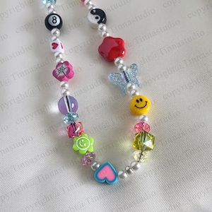 Pode incluir: Um colar de contas coloridas com uma variedade de pingentes, incluindo uma borboleta, um rosto sorridente, um cora&ccedil;&atilde;o, uma flor, uma estrela e um n&uacute;mero oito. O colar &eacute; feito com contas de p&eacute;rolas brancas e contas de metal prateado.