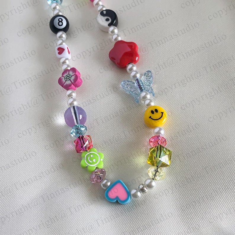 Pode incluir: Um colar de contas coloridas com uma variedade de pingentes, incluindo uma borboleta, um rosto sorridente, um cora&ccedil;&atilde;o, uma flor, uma estrela e um n&uacute;mero oito. O colar &eacute; feito com contas de p&eacute;rolas brancas e contas de metal prateado.
