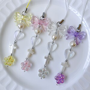 May include: Four colorful phone charms with a bow, heart, cross, and flower design. The charms are made of clear plastic with iridescent accents. The bows are in yellow, pink, clear, and purple.