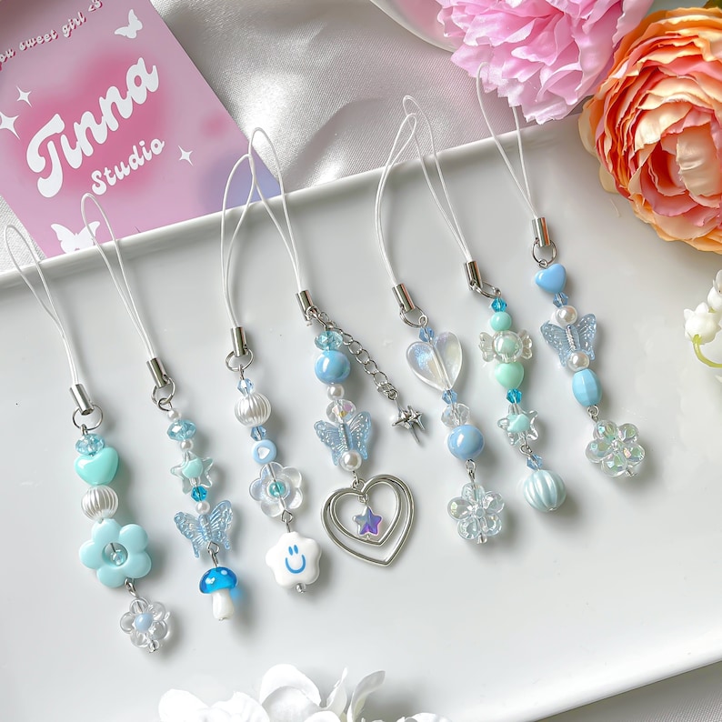 May include: Seven blue and white phone charms with different designs, including butterflies, hearts, stars, and flowers. The charms are on a white background with a pink card that says "Tinna Studio" in the background.