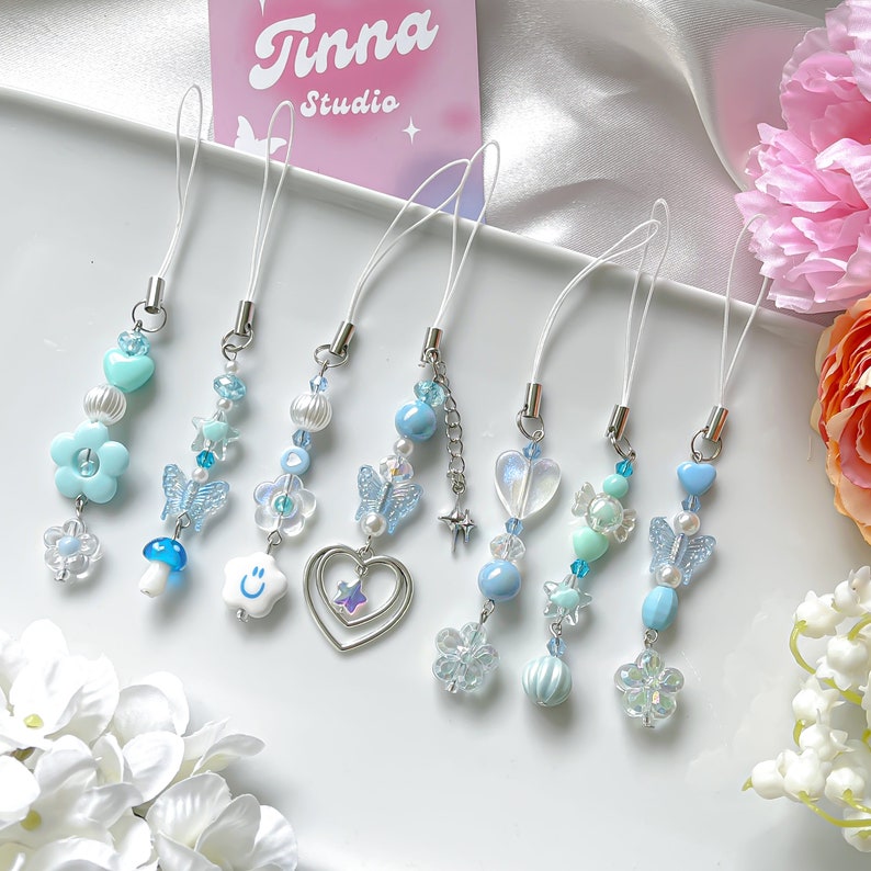 May include: Six blue and white beaded phone charms with different designs, including a heart, a butterfly, a flower, a mushroom, a smiley face, and a candy cane. The charms are hanging from white strings with silver clasps.
