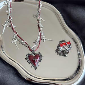 Op de afbeelding: Een zilverkleurige ketting met een rode hartvormige hanger en kruisdetail, gecombineerd met een bijpassende ring met een rode rechthoekige steen. De ketting bevat prikkeldraad- en kruisaccenten. De sieradenset wordt gepresenteerd op een zilveren dienblad.