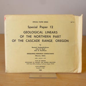 Op de afbeelding: Een vintage geologisch onderzoekspapier getiteld "Special Paper 12 Geological Linears of the Northern Part of the Cascade Range, Oregon". Het papier is vergeeld en heeft zwarte tekst. Het papier ligt op een houten oppervlak.