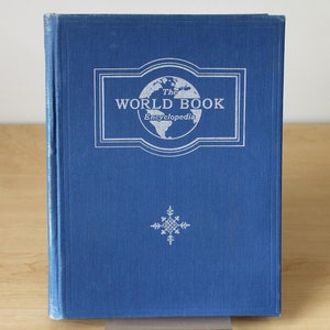 May include: A blue hardcover book with the title "The World Book Encyclopedia" in white lettering on the front cover. A white snowflake design is below the title.