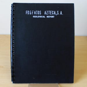 May include: A black bound report titled "FOSFATOS AZTECA, S.A. GEOLOGICAL REPORT" is displayed on a clear acrylic stand. The report has white text and a spiral binding on the left side. The background is a light-colored wooden surface.