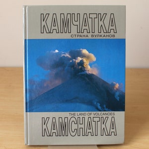 May include: A book titled "Kamchatka: The Land of Volcanoes" with a photo of a volcano erupting on the cover. The book cover is silver with black text.