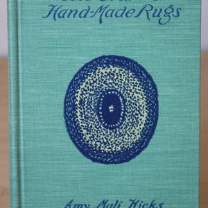 May include: A teal blue book with the title "The Craft of Hand-Made Rugs" in dark blue lettering. The cover features a blue and white circular design with a smaller blue circle in the center.