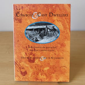 May include: A book cover with a brown and orange background. The title is "Cowboys & Cave Dwellers: Basketmaker Archaeology in Utah's Grand Gulch" by Fred M. Blackburn & Ray A. Williamson. The cover features a black and white photo of a group of people digging in a dirt pit.