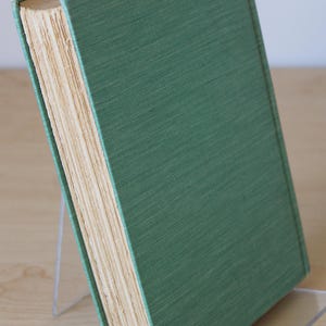 May include: A green hardcover book with a spine that is slightly worn. The book is standing upright on a clear acrylic bookend.