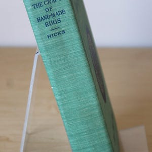 May include: A green book titled "The Craft of Hand-Made Rugs" by Hicks. The book is on a white stand and has the word "Empire" printed on the bottom.
