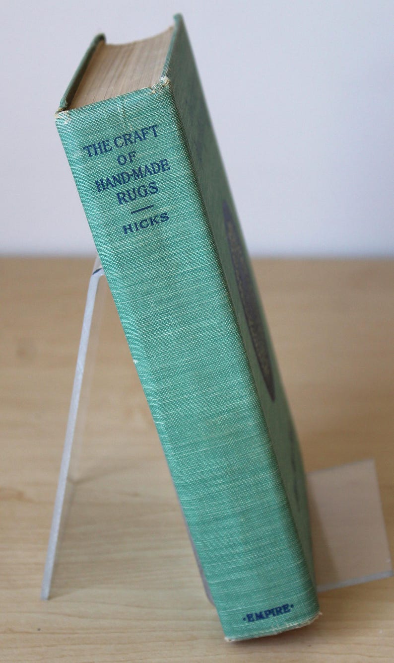 May include: A green book titled "The Craft of Hand-Made Rugs" by Hicks. The book is on a white stand and has the word "Empire" printed on the bottom.