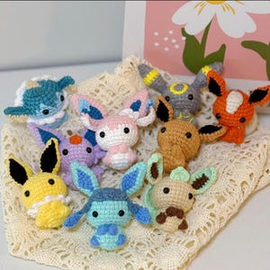 May include: A collection of handmade crochet plush toys in various colors, including blue, pink, yellow, and orange. The toys have large ears and are arranged on a cream-colored, open-weave fabric. Each toy has black eyes and unique features.