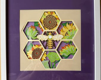 May include: A paper cut-out art piece depicting a bee in the center of a honeycomb with six sunflowers surrounding it. The honeycomb and sunflowers are made of paper and are layered to create a 3D effect. The bee is yellow and black with white wings. The sunflowers are yellow with brown centers. The background is purple.