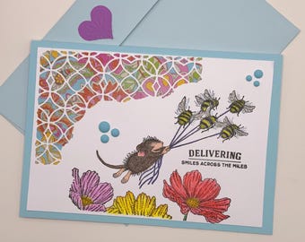 May include: A greeting card with a mouse holding a bunch of bees, with the text "Delivering Smiles Across The Miles". The card has a colorful patterned background and flowers.