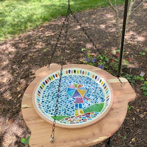 May include: A mosaic bird bath with a colorful umbrella design hangs from a black metal stand. The bath is made of small, colorful tiles and is surrounded by a wooden platform.