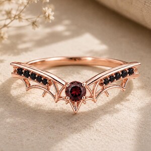May include: A rose gold ring featuring a V-shaped band with a central round-cut red garnet gemstone. The band is accented with small black stones along each side, and the metalwork is shaped to resemble bat wings extending from the center stone.