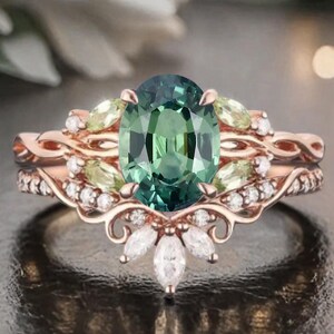 May include: A rose gold engagement ring set with an oval, teal-green gemstone as the centerpiece. The ring is flanked by smaller green gemstones and white diamonds. The band features a delicate, floral design. A matching band sits below the engagement ring.