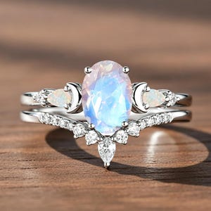 May include: A silver ring set featuring a large, oval-cut, iridescent gemstone as the centerpiece. The ring is accompanied by a matching band with a curved design, adorned with smaller gemstones and moon-shaped accents. The set is displayed on a wooden surface.