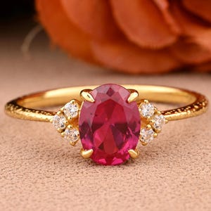 May include: A gold ring featuring an oval-cut, ruby-red gemstone as the centerpiece. The ring is accented with small, round, clear gemstones on each side of the main stone. The band has a textured design.