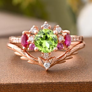 May include: A rose gold ring set featuring a peridot center stone, surrounded by smaller pink and white gemstones. The band has a leaf-like design. The ring set is on a brown surface.