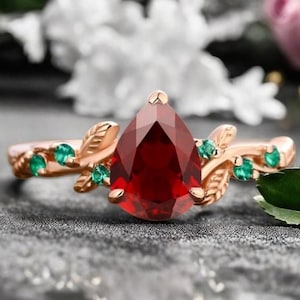 May include: A pear-shaped red gemstone ring with a gold band and green leaves. The ring is set on a gray background.