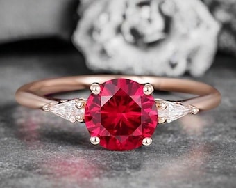 May include: A rose gold ring with a large, round, faceted red gemstone as the centrepiece. Two smaller, triangular, white gemstones flank the main stone. The ring is set against a grey background.